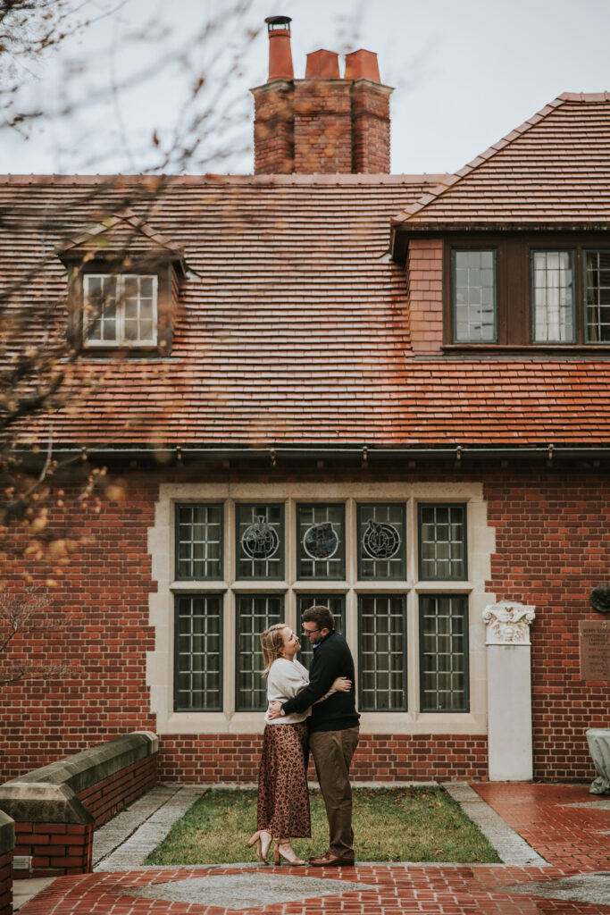 Couple standing together in front of Cranbrook House with colorful leaves around them.