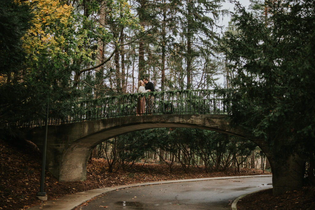 Couple posing on a bridge surrounded by vibrant fall foliage at Cranbrook House and Gardens.