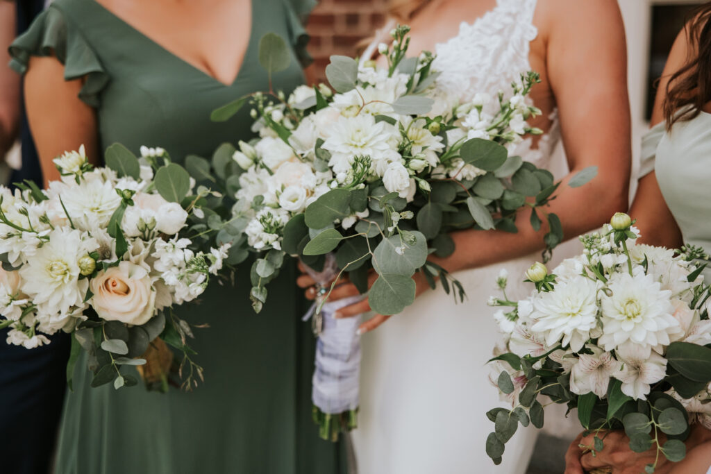 Close-up of bride’s wedding bouquet, featuring white flowers and greenery with Lovett Hall in the background.