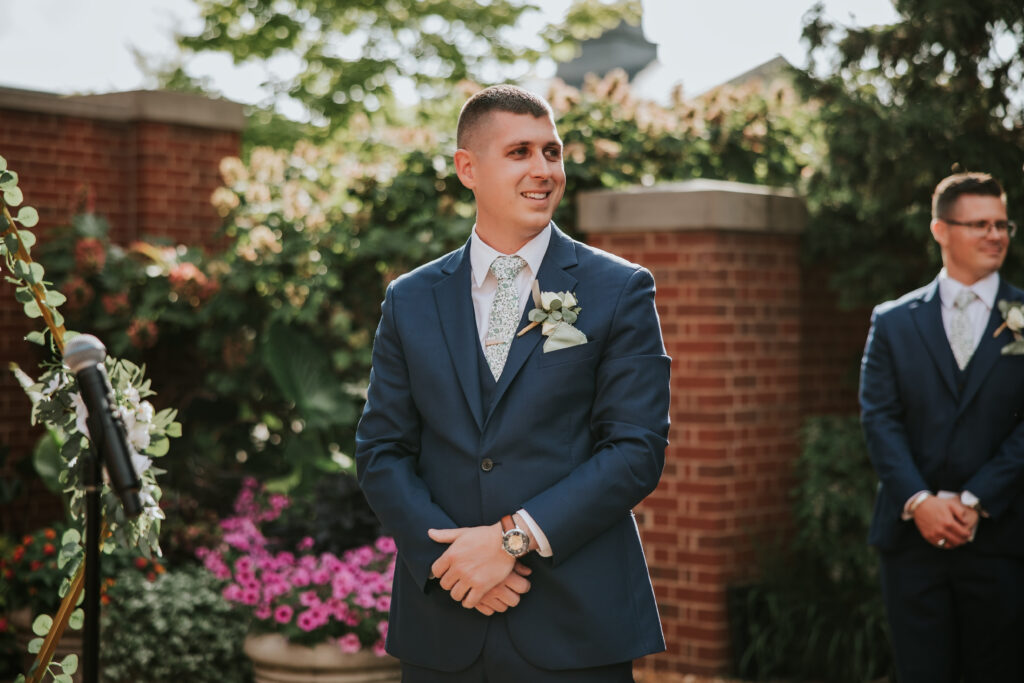 Groom smiling with joy as he sees his bride as she walks down the aisle at the Ginger Meyer Garden outside of Lovett Hall.