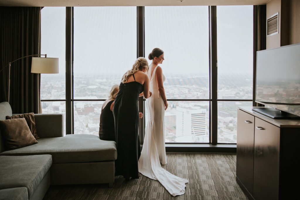 Bride getting ready by a large window, with bridesmaids buttoning her wedding dress | Wedding Planning Tips